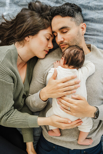 new parents snuggle together on bed with newborn baby on chest