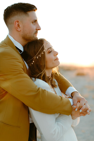 groom holding his bride from behind and clasping hands in the front. both are looking off with a soft smile at the sunset.