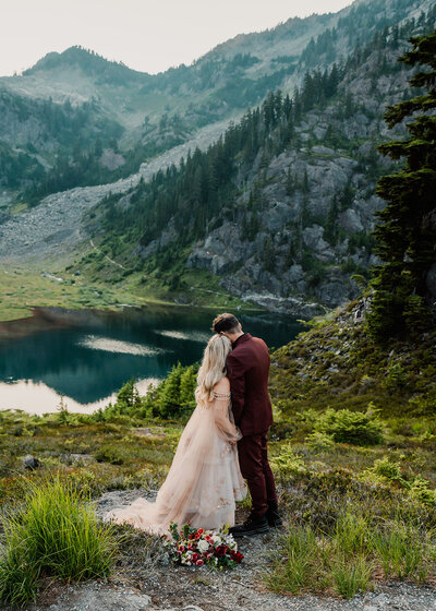 a couple in wedding attire embrace in a stunning blue and green landscape. Their adventure elopement photographers, shoot from a higher vantage point showing off the mountains and alpine lake the couple is gazing at. 
