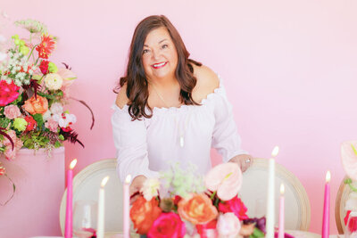 Susan Noland in light pink top smiling while adjusting a floral centerpiece on a table in front of a pink wall