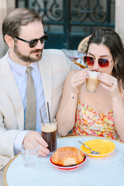 Couple at a Parisian café in the Marais near Hôtel de Sully, wearing sunglasses as the woman sips coffee during a couple’s photoshoot in Paris.