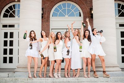 Senior girls wearing white dresses standing on steps of Wyatt Center popping champagne and smiling in celebration of graduation