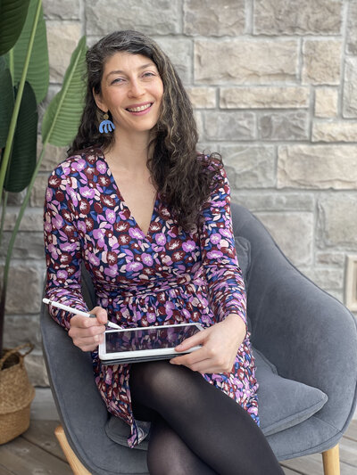 A portrait of artist and textile designer Skye McNeill. Skye wears a blue cardigan and a black shirt and stands in front of a wall sized block print of palm leaves. Photograph by Beth Olsen Creative.