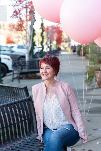 Woman sitting on a bench holding  pink balloons and smiling.