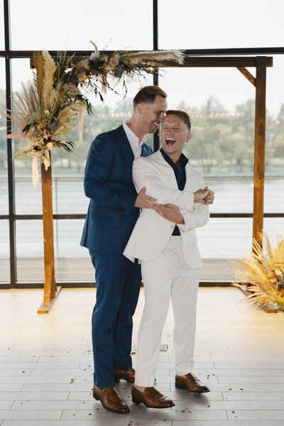 Two grooms standing one in front of the other holding each other and laughing 