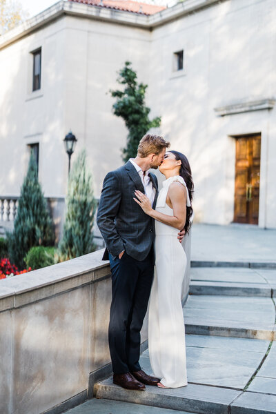 Man wearing suit kissing woman wearing white pant suit on University of Pittsburgh campus