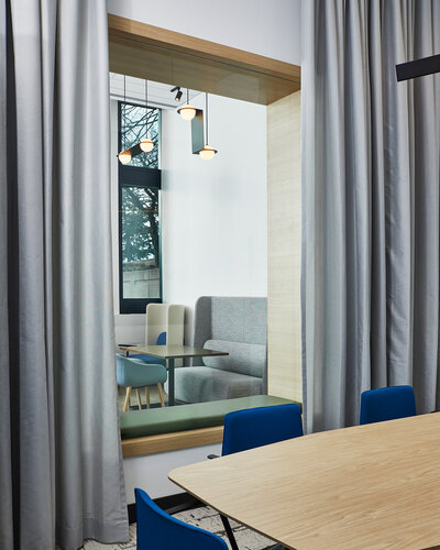 A wood table surrounded by blue chairs, framed by off-white curtains and overlooking another bright meeting area