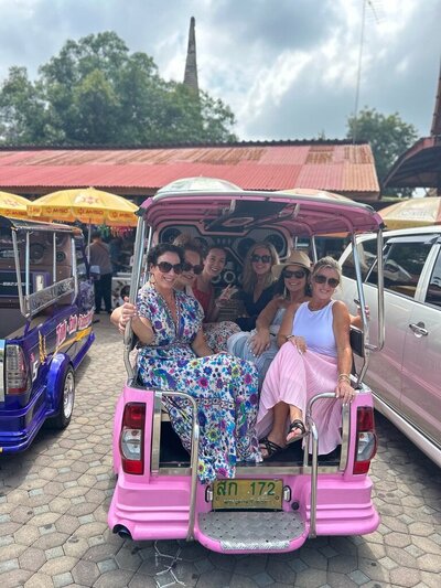 A group of six women, all smiling, sit closely together in a bright pink tuk-tuk, enjoying a fun ride. The women are dressed in colorful, casual clothes and sunglasses, with a vibrant market scene in the background.