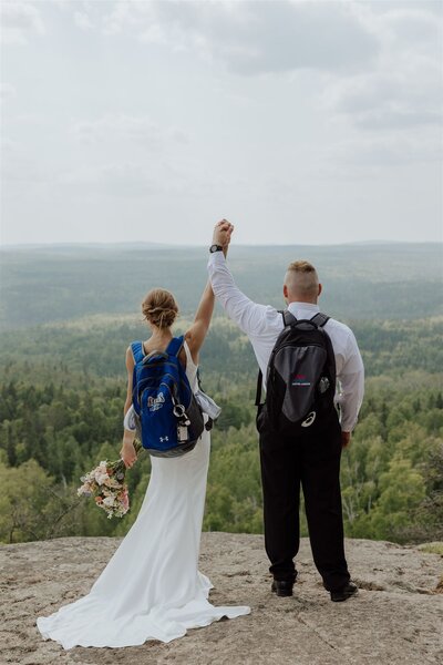 bride and groom wearing hiking backpacks holding hands and celebrating on a rocky overlook. captured by a minnesota elopement photographer