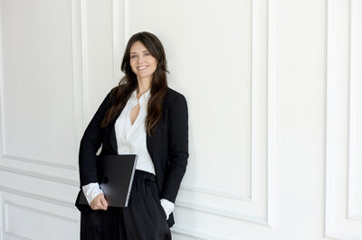 woman in a black suit leaning against a french wall in a black suit holding a laptop smiling at the camera
