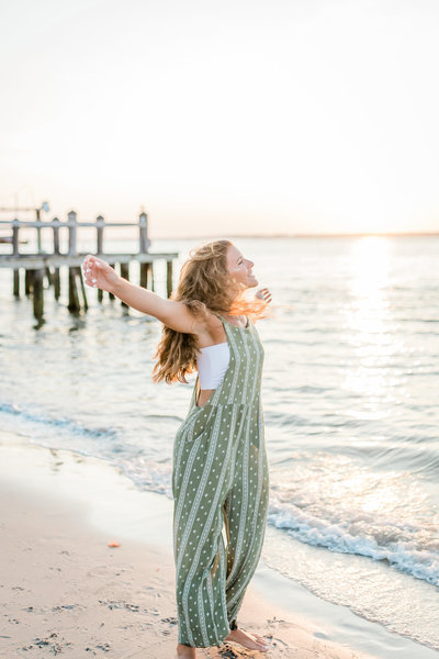 High school senior portrait of a girl playing in the sand on the beach in Ocean City, New Jersey. She is wearing a white romper and the sun is glowing.