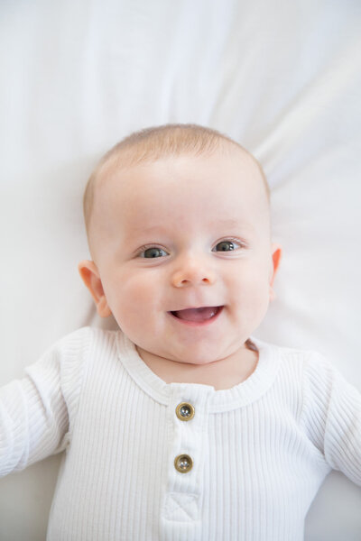 Happy infant boy in white on a white bed, captured by las vegas portrait photographer Jessica Bowles