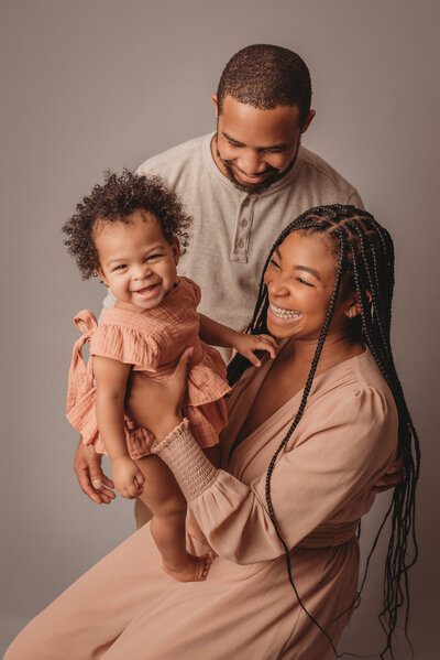 Family portrait with mom, dad smiling at one year old baby girl, who is smiling at the camera on gray backdrop