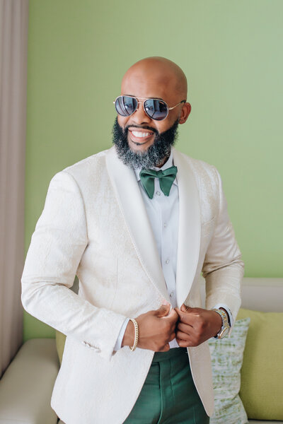 A groom smiles confidently while adjusting his white tuxedo jacket. He is dressed in a green bow tie and matching green pants, wearing sunglasses indoors against a light green wall