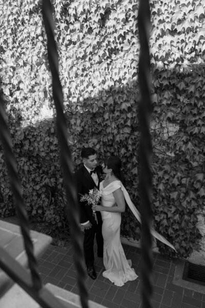 Through bars on a staircase you see the bride and groom standing together, in black and white.