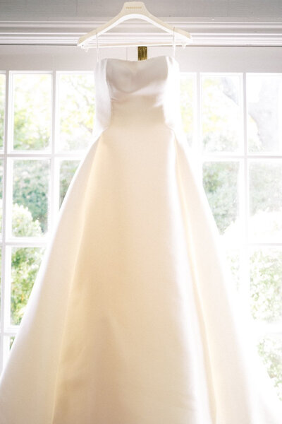 classic, simple satin wedding dress hanging in front of a bright window in a serene room