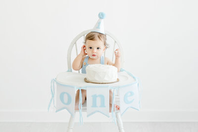 One year old baby boy sits in high chair wearing birthday hat for a cake smash portrait session
