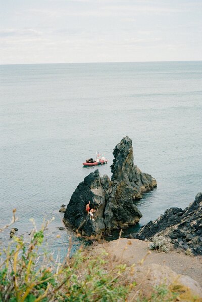 Two people climbing dark rock formations by the sea, with a red inflatable boat floating nearby in the water.