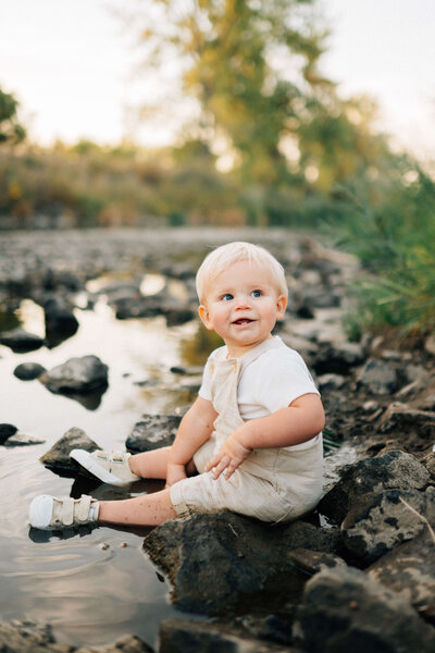 baby boy sitting on the riverbank playing with rocks and water