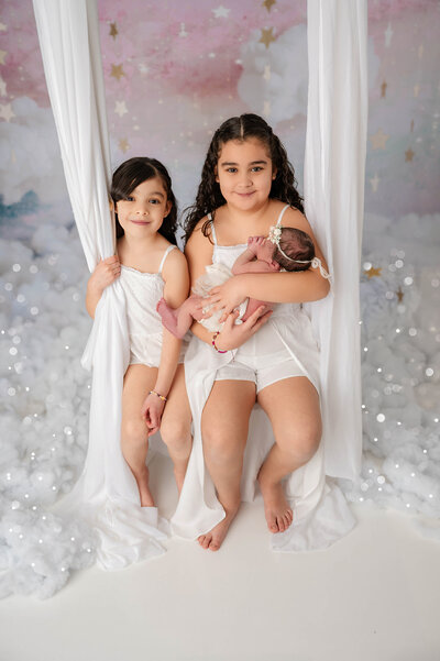 Two older girls sit on a white draped swing, the oldest cradling a newborn, with fluffy clouds and stars in the background.