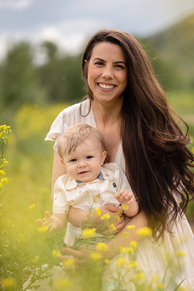 Bright and cheerful portrait of a mother with long brown hair, holding her baby boy amongst yellow flowers in Westsyde, Kamloops. Portrait by Brittany Danielle Photography
