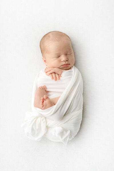 A newborn baby sleeps with their hands folded underneath their cheek. The baby is lightly swaddled in a creamy white blanket, with their folded feet peeking out from above the knot of the blanket. The newborn was photographed in studio by Karen Kahn of Looking Up Photography.