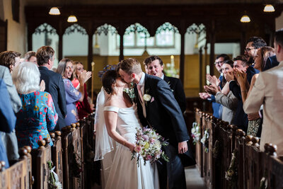 Bride and groom kiss in the middle of the church aisle after just marrying. The groomsman behind and guests either side are cheering and clapping. 