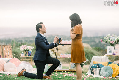 Future Groom is down one knee as he proposes to his girlfriend with their memories all around them at the Coastal Vista View Park