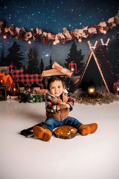 Baby boy in plaid flannel and work boots sits among honey pots at woodland themed first birthday portrait session