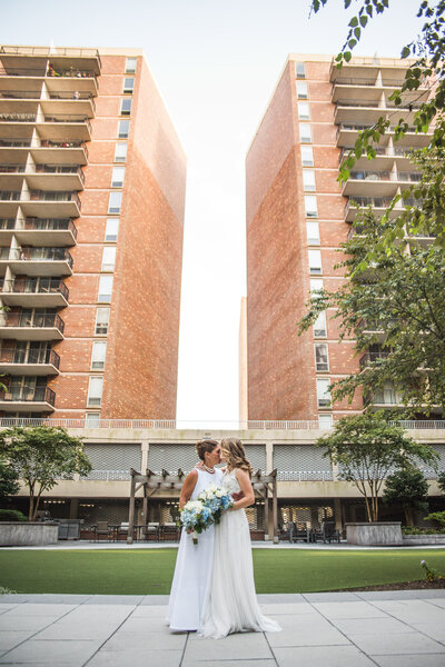 A newlywed couple kissing in front of two large brick buildings 