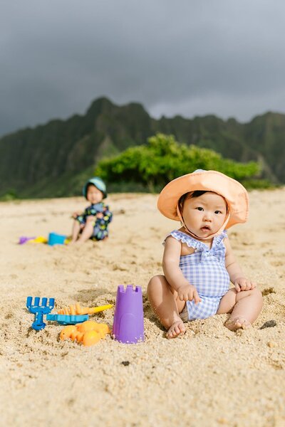 A baby, wearing a sun hat and a little boy in the background play in the sand on the beach at Kailua.