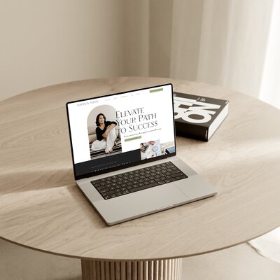 A laptop displaying a Showit template on a circular wooden table. A book titled "Do" is placed next to the laptop. The background shows a white curtain, creating an inspiring workspace for women entrepreneurs seeking sophisticated website design services.