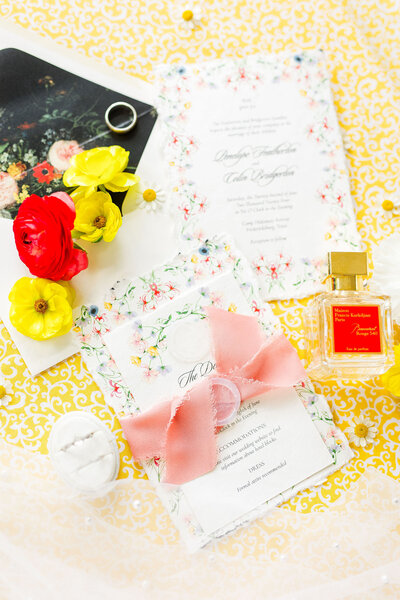 Flat lay with a yellow background, perfume, florals, rings, and wedding stationary