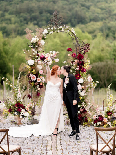 A couple kiss in front of a flower arch at a Chateau St Agnes wedding ceremony.