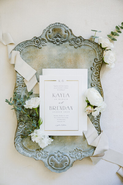A beautifully styled wedding invitation suite displayed on an ornate antique silver tray, adorned with soft silk ribbons, white florals, and delicate greenery. Captured at The Gibbes Museum in Charleston, SC, this romantic detail shot exudes timeless elegance and luxury.