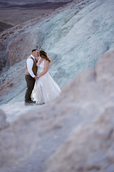 A couple in wedding clothes kiss while standing among the pastel colors of Artists Palette in Death Valley