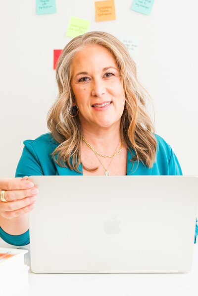 Woman in teal blazer opening an apple macbook. Wall beind her has sticky notes with inspirational phrases on them.