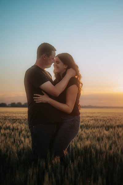 A couple stands embracing in a wheat field at sunset.