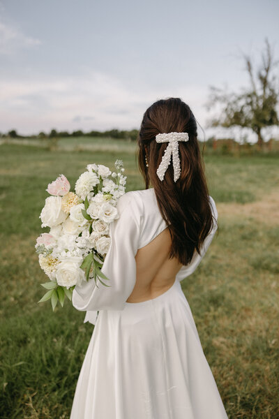Bride standing in an open grassy field holding a lush white and soft-blush garden-style elopement bouquet featuring roses, lisianthus, dahlias, and delicate greenery; wearing a long-sleeve open-back wedding dress with a pearl bow hair accessory, captured from behind during golden-hour portraits.