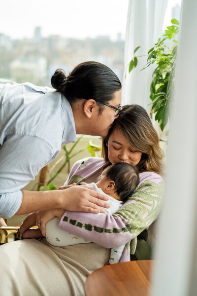A father leans in to kiss his wife as she kisses their sleeping baby boy. A romantic moment captured at their in-home newborn photo shoot for their older baby. 