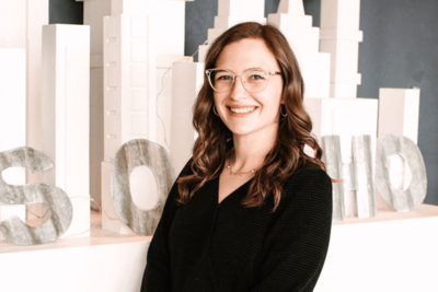 Kalie Breeze, general manager of SoHo63, a Chandler wedding and event venue, smiles while standing in front of a modern white cityscape backdrop. Her professional and friendly demeanor reflects the exceptional service and hospitality SoHo63 is known for.