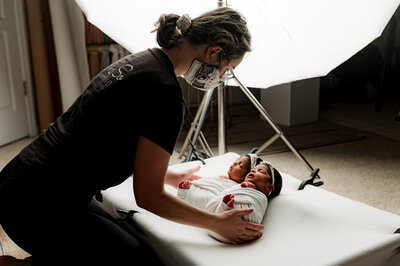 Sofia Patrao Charlotte newborn photographer at NicSo Studio, wearing a mask gently arranges twin sleeping newborns wrapped in blankets on a cushioned surface. Professional lighting in the background hints at an enchanting photography session.