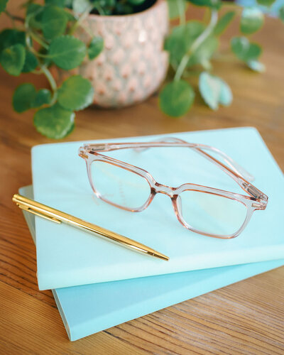 Pink reading glasses and a gold pen sit  on top of two blue notebooks as a representation of brand imagery for small business copywriting services.