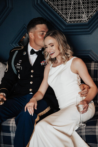 Groom in uniform kissing bride on head. Sitting on couch with glass of champagne in hands