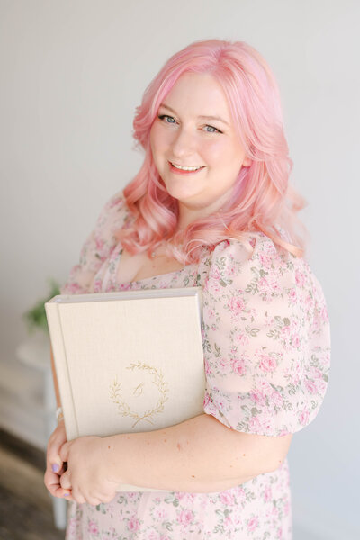 Woman with pink hair in a pink floral dress holds an heirloom album and smiles, Indianapolis Photographer Brittney Lear