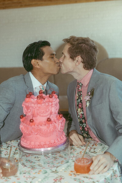 Two grooms kissing with a wedding cake in front of them.