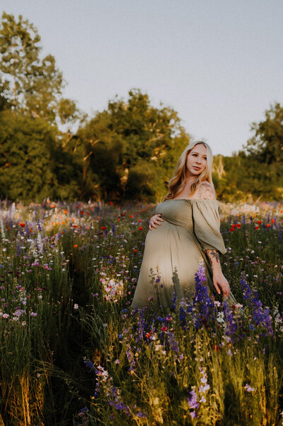 A pregnant mom wearing a sage green dress holding her belly and walking in a field of wildflowers in Richardson, Texas
