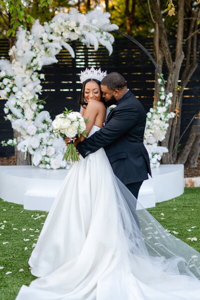 Groom embraces his bride while she holds her bouquet in front of floral installation