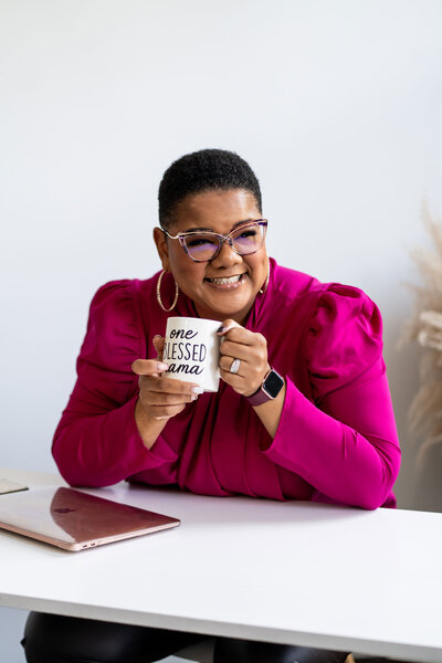Career strategist expert working on her laptop at a white office desk, dressed in a professional branded pink outfit, holding a coffee cup with an inspirational quote.