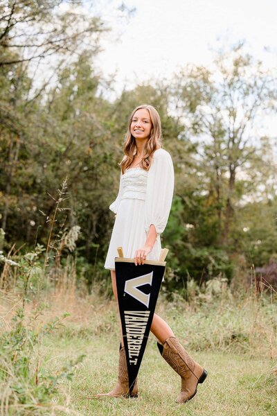 ardrey kell senior stands in field hoding vanderbilt pennant wearing a cream dress and boots